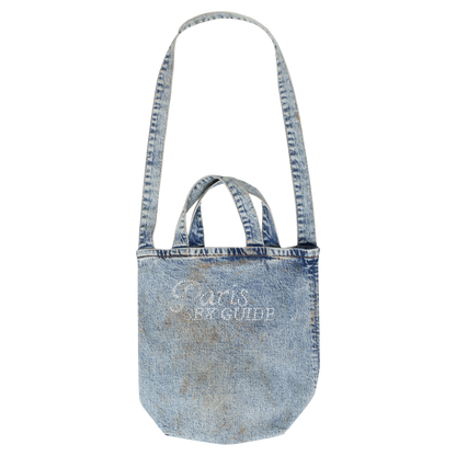 Freigestellte Umhängetasche aus hellem, ausgewaschenem Denim mit sichtbaren Tragespuren. Die Tasche verfügt über zwei kurze Tragehenkel sowie einen langen Schulterriemen. Auf der Vorderseite befindet sich ein weißer Strass-Schriftzug mit der Aufschrift „Paris SEX GUIDE“. Die Form ist leicht abgerundet mit weichem Boden.